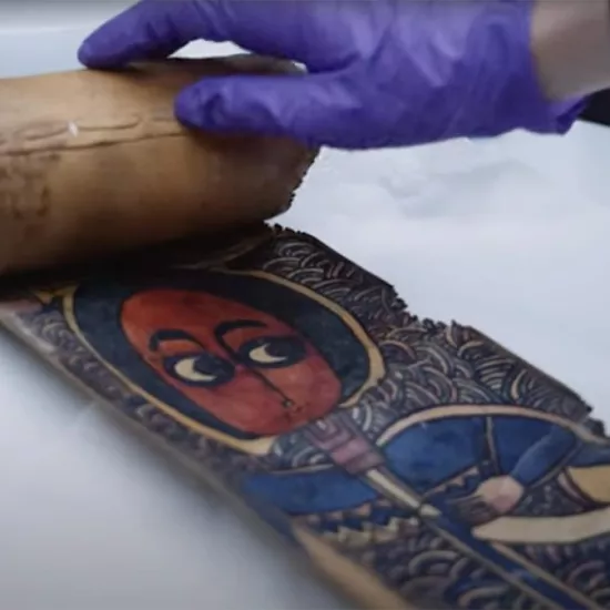Hand wearing latex-like glove unrolling a scroll about a hands-width with colourful image of a person with round head, large eyes, and a blue robe 