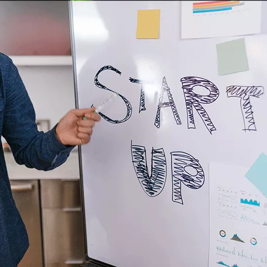 Hand holding a marker points to the words "Start Up" written on a whiteboard.