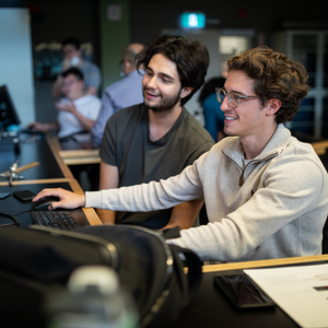Two male students talk and laugh while working at a computer.