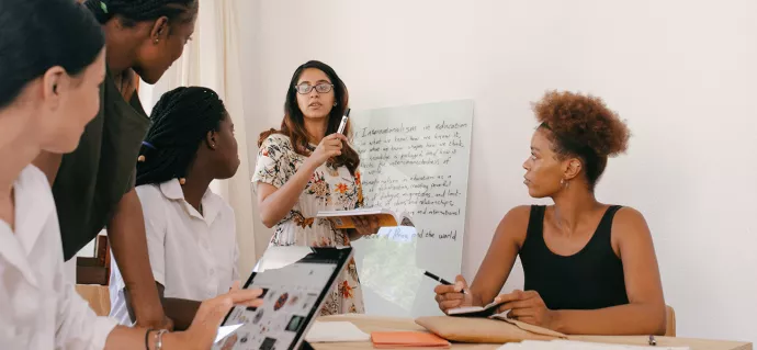 A woman runs a meeting, writing on a whiteboard while four colleagues take notes.