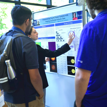 A female researcher points to her poster and explains her research as students listen.